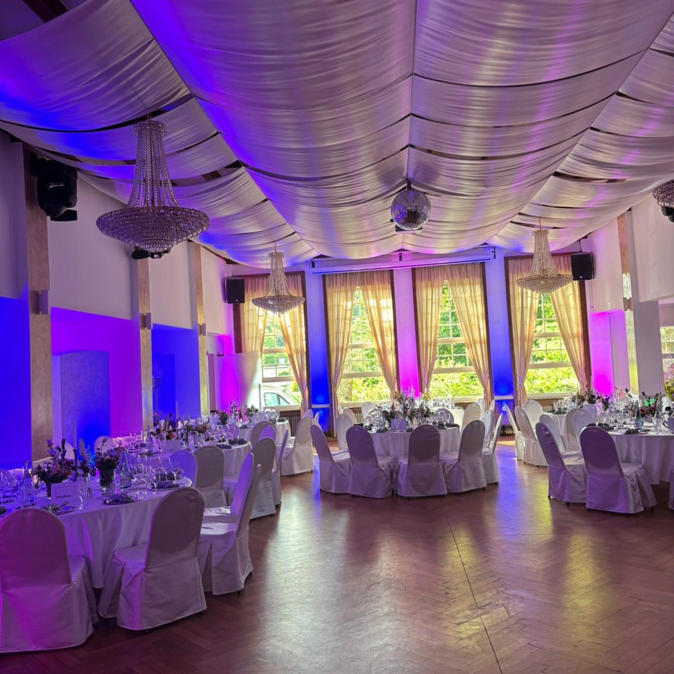 Ambientebeleuchtung für Hochzeit im Münsterland Eleganter Veranstaltungsraum mit runden Tischen, weißen Stühlen und lila Beleuchtung.