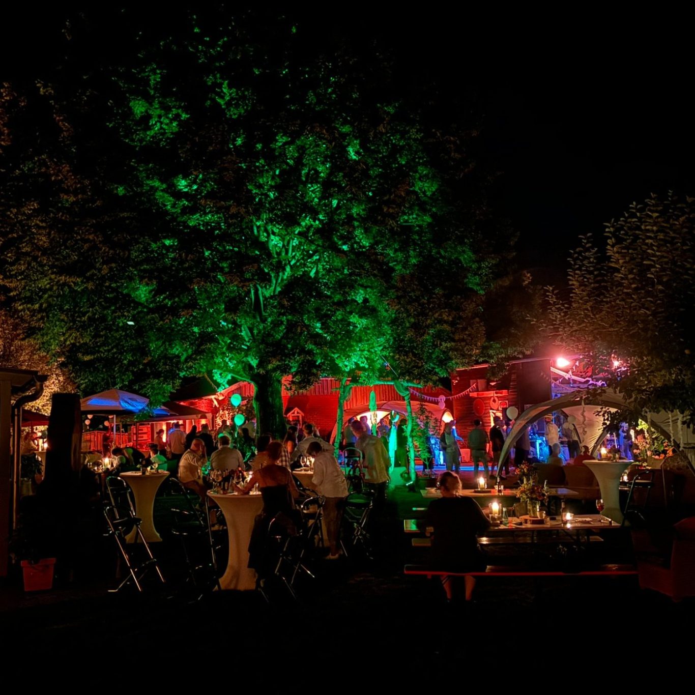 Stimmungsvolle Beleuchtung für Greven und das Münsterland Bunt beleuchteter Garten mit Menschen, Sitzgelegenheiten und Bäumen bei Nacht.